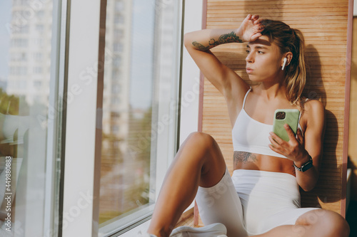 Young tired skinny sportswoman woman in white sportswear earphones listen music use mobile cell phone put hand forehead breathing sit on windowsill of gym rest indoors People urban lifestyle concept Free Download Young tired skinny sportswoman woman in white sportswear earphones listen music use mobile cell phone put hand forehead breathing sit on windowsill of gym rest indoors People urban lifestyle concept - GFXLoad