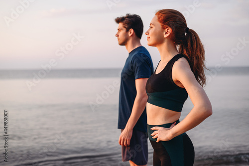 Side view couple young two friend strong sporty sportswoman sportsman woman man in sport clothes warm up train stand akimbo do exercise on sand sea ocean beach outside on seaside in summer day morning