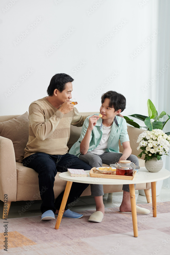 Joyful mature man enjoying spending weekend with son at home, they are drinking tea, eating potato chips and discussing news