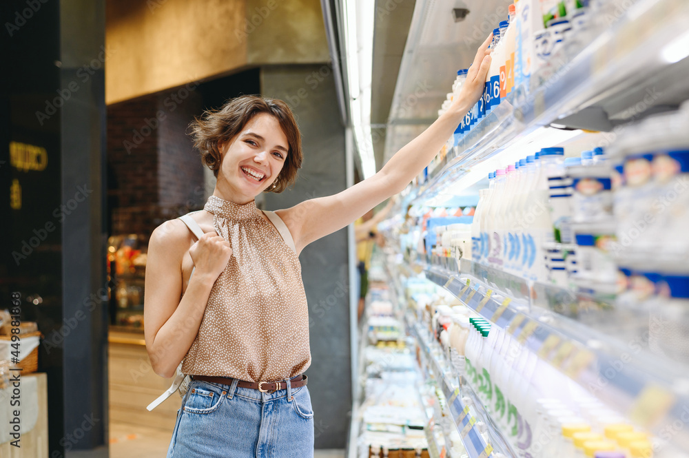 Young smiling happy fun woman 20s wear casual clothes backpack shopping at supermaket store buy choose dairy produce taking milk bottle inside hypermarket. People purchasing gastronomy food concept.