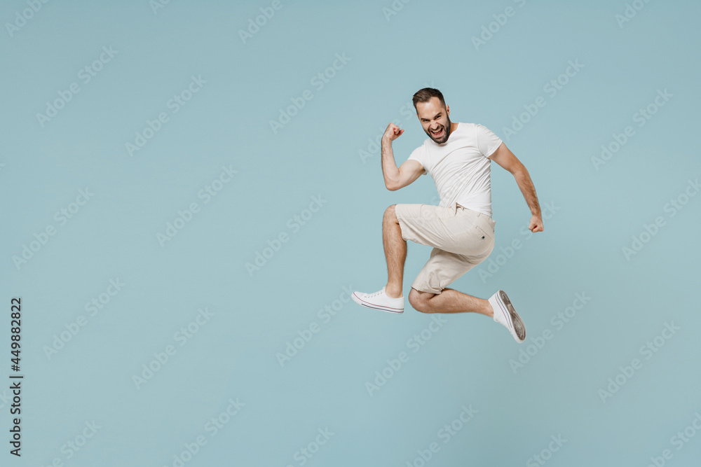 Full length side view young overjoyed happy excited man 20s wearing casual white t-shirt jump high do winner gesture clench fist isolated on plain pastel light blue color background studio portrait