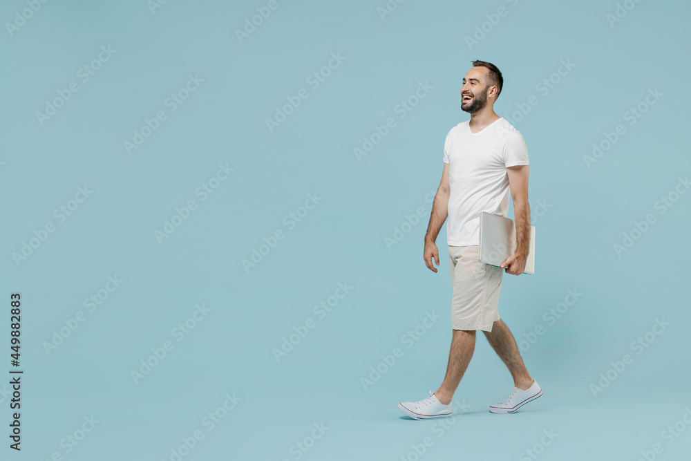 Full length side view young confident smiling happy minded man 20s in white t-shirt walking going hold closed laptop pc computer look aside isolated on plain pastel light blue color background studio Full length side view young confident smiling happy minded man 20s in white t-shirt walking going hold closed laptop pc computer look aside isolated on plain pastel light blue color background studio