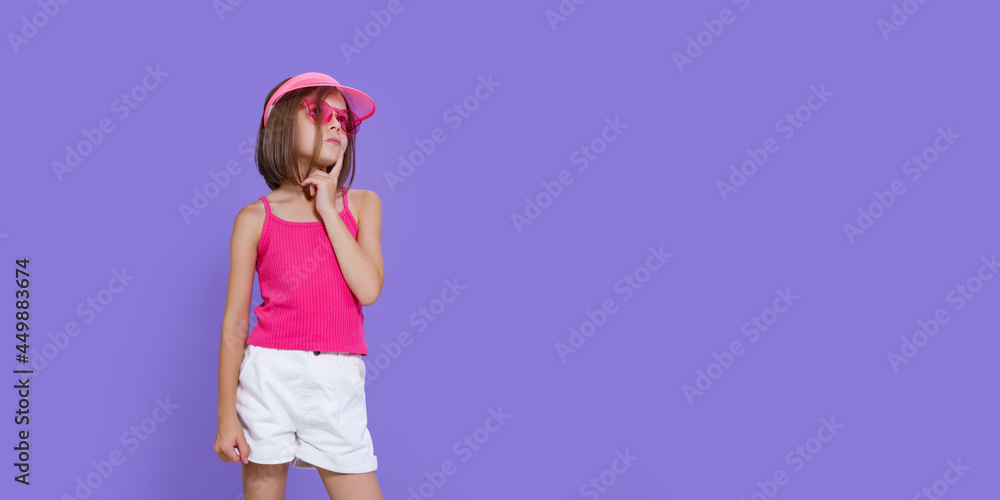 Little thoughtful girl in white shorts, a pink T-shirt, pink trendy glasses and a summer visor on a purple background in the studio Little thoughtful girl in white shorts, a pink T-shirt, pink trendy glasses and a summer visor on a purple background in the studio
