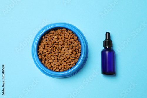 Glass bottle of tincture near bowl with dry pet food on light blue background, flat lay