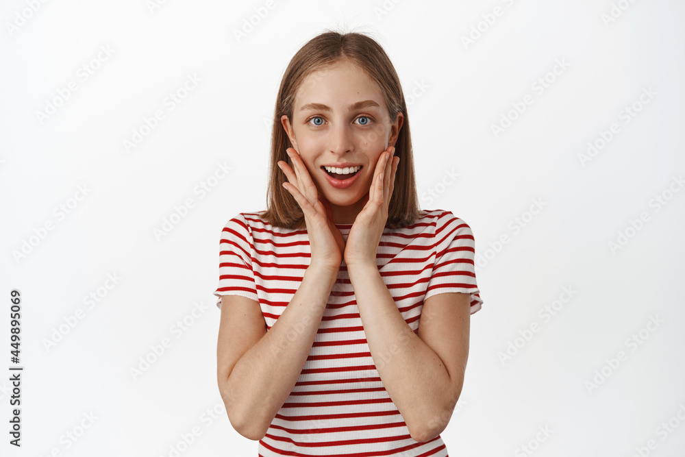 Portrait of delighted smiling blond girl feel flattered and touched, looking surprised at gift, hold hands near face and gazing at camera in awe, standing against white background Portrait of delighted smiling blond girl feel flattered and touched, looking surprised at gift, hold hands near face and gazing at camera in awe, standing against white background
