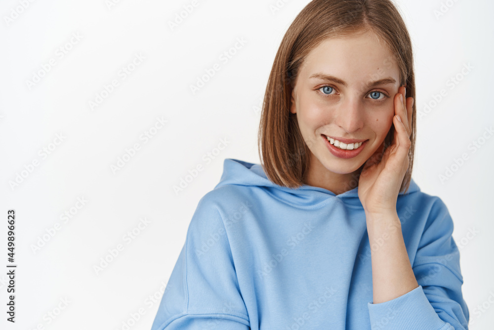 Women face and skin care. Portrait of beautiful blond girl touching her cheek, smiling and gazing happy at camera, dont be shy drop your cosmetics brand name, white background