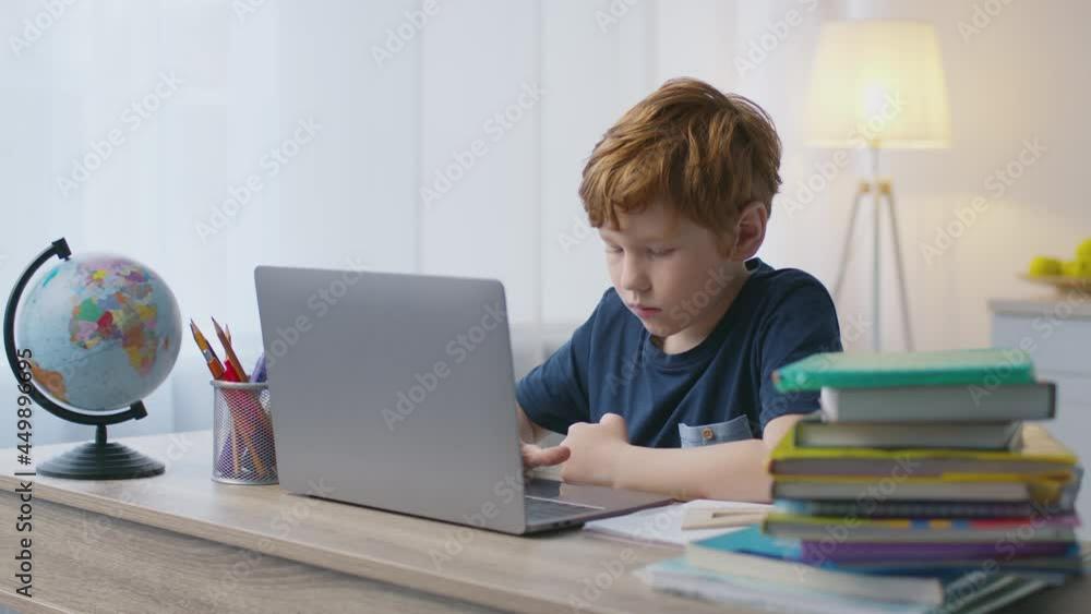Homeschooling concept. Cute little boy typing on laptop computer, sitting at home workplace with books and globe Homeschooling concept. Cute little boy typing on laptop computer, sitting at home workplace with books and globe