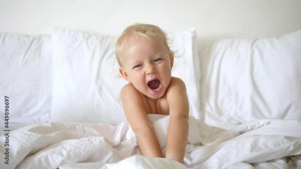 Portrait of cute 2 years old caucasian baby sitting in an adult white bed yawning and throwing himself onto bed Portrait of cute 2 years old caucasian baby sitting in an adult white bed yawning and throwing himself onto bed
