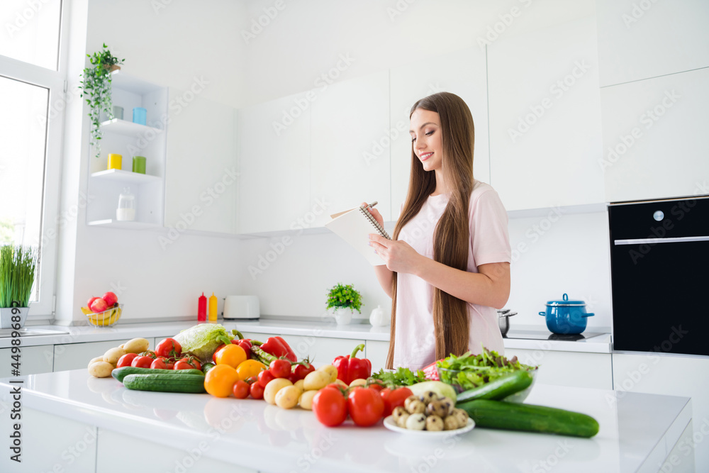 Portrait of attractive cheerful long-haired girl cooking useful salad farm product writing plan list at home light white kitchen indoors