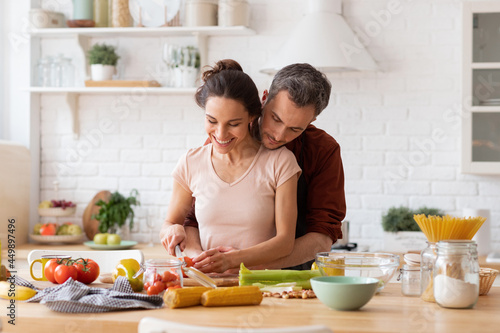 Happy wife and husband cooking romantic dinner at home kitchen. Man hugging woman from back. Couple chopping vegetable together. Fresh salad recipe preparation. Loving relationship. Family portrait