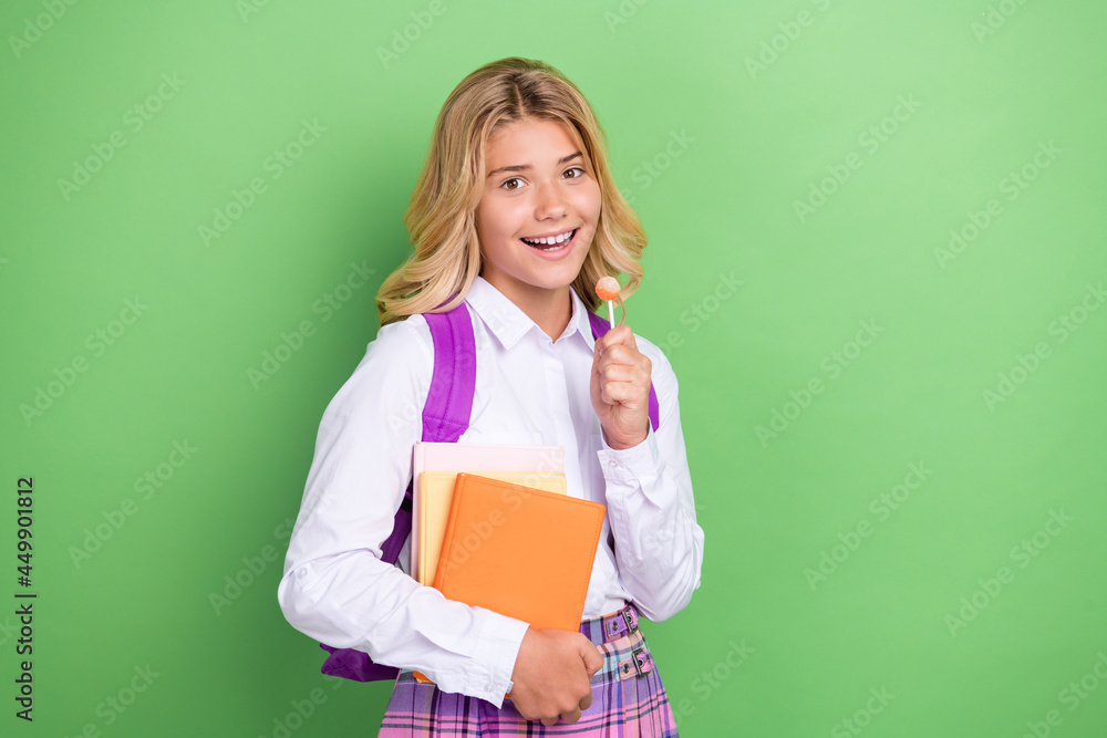Portrait of attractive cheerful funny girl eating caramel holding book going to lesson isolated over green color background Portrait of attractive cheerful funny girl eating caramel holding book going to lesson isolated over green color background