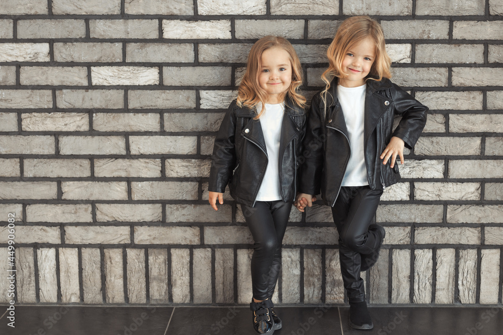 Street style portrait of two cute pretty sisters 2-3 years old in same outfits: white t-shirts, leather black jackets and leggings. Best friends together outdoors. Street style portrait of two cute pretty sisters 2-3 years old in same outfits: white t-shirts, leather black jackets and leggings. Best friends together outdoors.