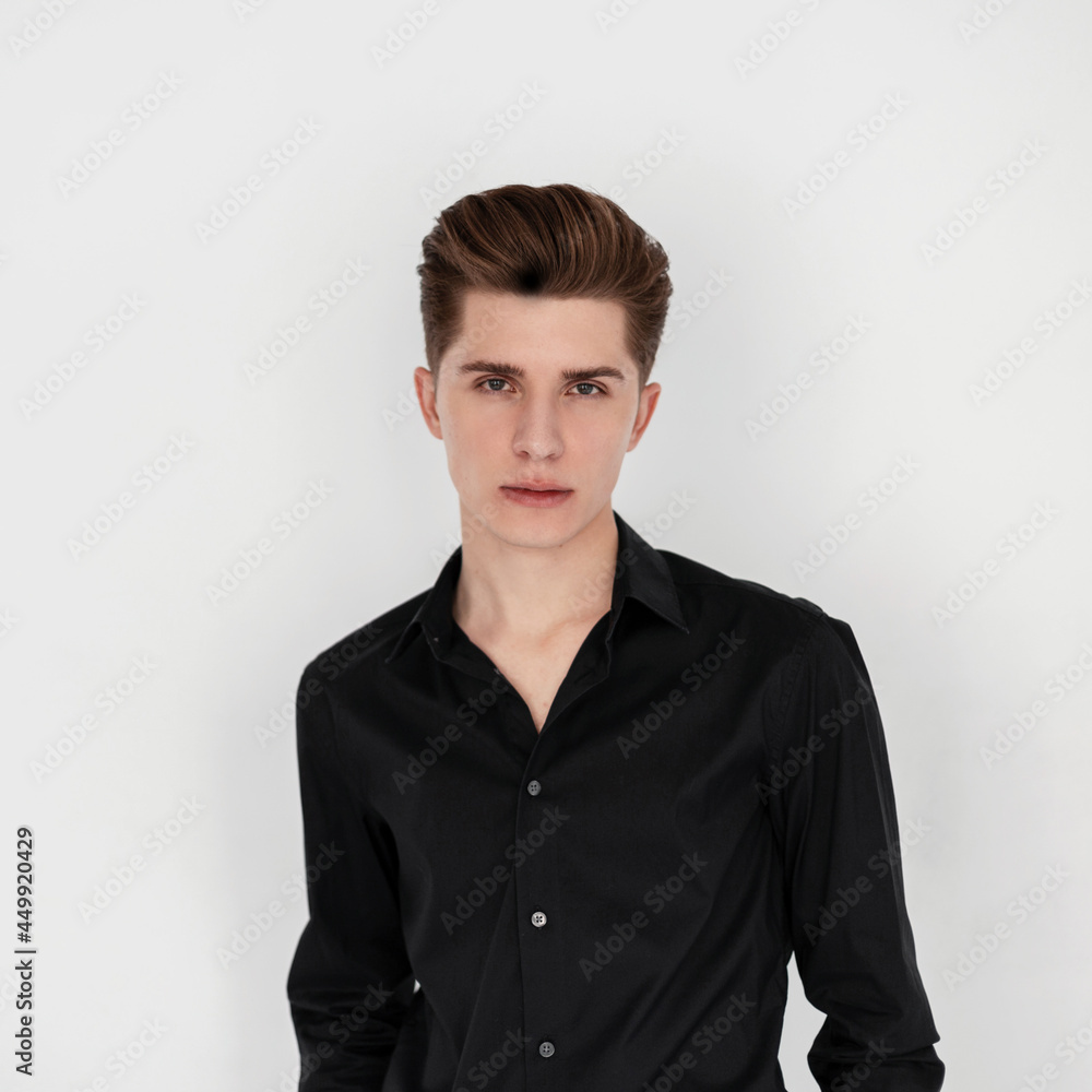 Portrait stylish handsome young man in elegant trendy black shirt on white background indoors. Attractive guy fashion model with trendy hairstyle stands near vintage wall in studio. Beauty menswear. Portrait stylish handsome young man in elegant trendy black shirt on white background indoors. Attractive guy fashion model with trendy hairstyle stands near vintage wall in studio. Beauty menswear.