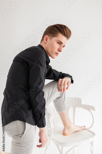 European elegant young man in fashion black shirt in in fashionable pants stands near wooden chair in studio. Handsome guy model in classic stylish clothes near white wall in room. Trendy menswear.