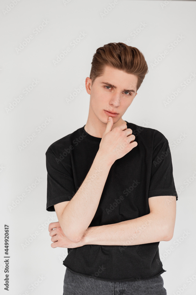 Pretty attractive young man fashion model with hairstyle in black stylish casual t-shirt in vintage jeans posing on a white background in studio. Fresh fashionable portrait european handsome guy. Pretty attractive young man fashion model with hairstyle in black stylish casual t-shirt in vintage jeans posing on a white background in studio. Fresh fashionable portrait european handsome guy.