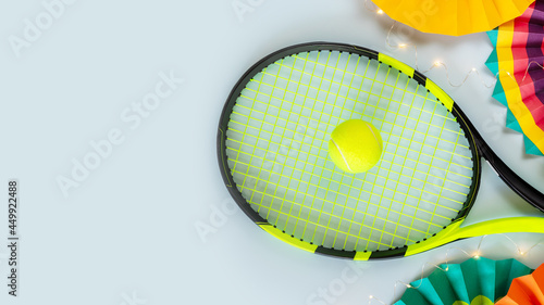 Tennis, horizontal composition with racket, yellow tennis ball, colorful paper fans and a light garland on a blue background with place for text. Holiday banner for a tennis competition. Flat lay