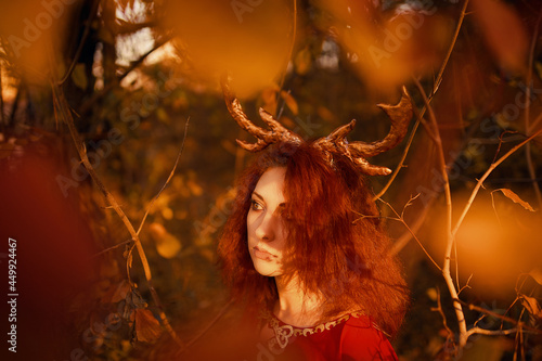 Woman in long red dress with deer horns in autumn forest. The demon of fairy tales in a red cloak with a lantern in his hands walks through the autumn forest and looks for magic herbs.