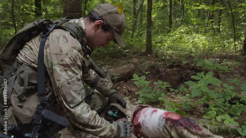 Close-up of combat medic taking first aid kit, providing emergency medical treatment to injured soldier with bandaged wounded leg. Healthcare Specialist providing emergency assistance to warrior Close-up of combat medic taking first aid kit, providing emergency medical treatment to injured soldier with bandaged wounded leg. Healthcare Specialist providing emergency assistance to warrior