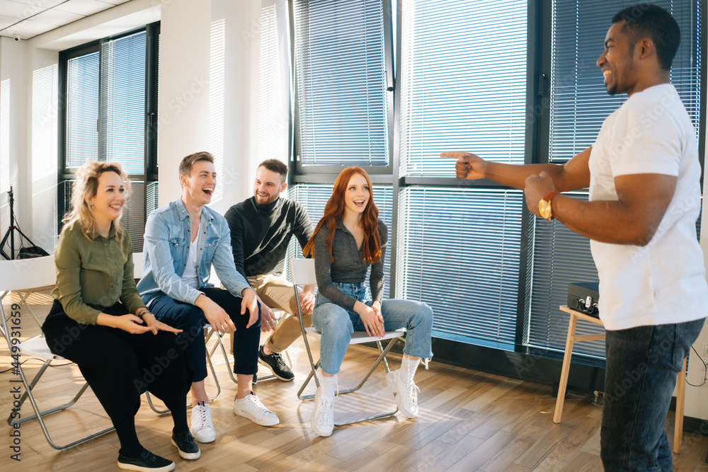 Group of young diverse multiethnic colleagues playing in active games during team building in modern office room. Cheerful African American male playing charades with friends showing pantomime.
