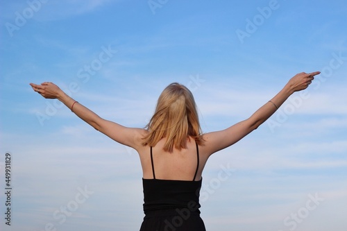 A young caucasian woman with her hands up in a black T-shirt against a blue sky. The concept of freedom, happiness, success, health. Back view