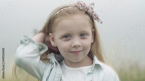 Headshot of pretty Caucasian little girl touching long hair looking at camera standing outdoors on foggy misty day. Close-up portrait of smiling charming beautiful child posing on meadow countryside photo