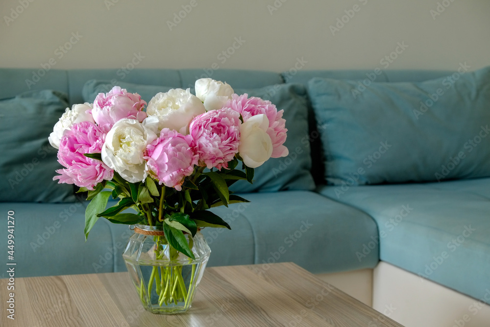 Close up shot of wooden coffee table with bouquet of beautiful white peony flowers in glass vase on foreground and blue textile couch on the background. Copy space for text, close up. Close up shot of wooden coffee table with bouquet of beautiful white peony flowers in glass vase on foreground and blue textile couch on the background. Copy space for text, close up.
