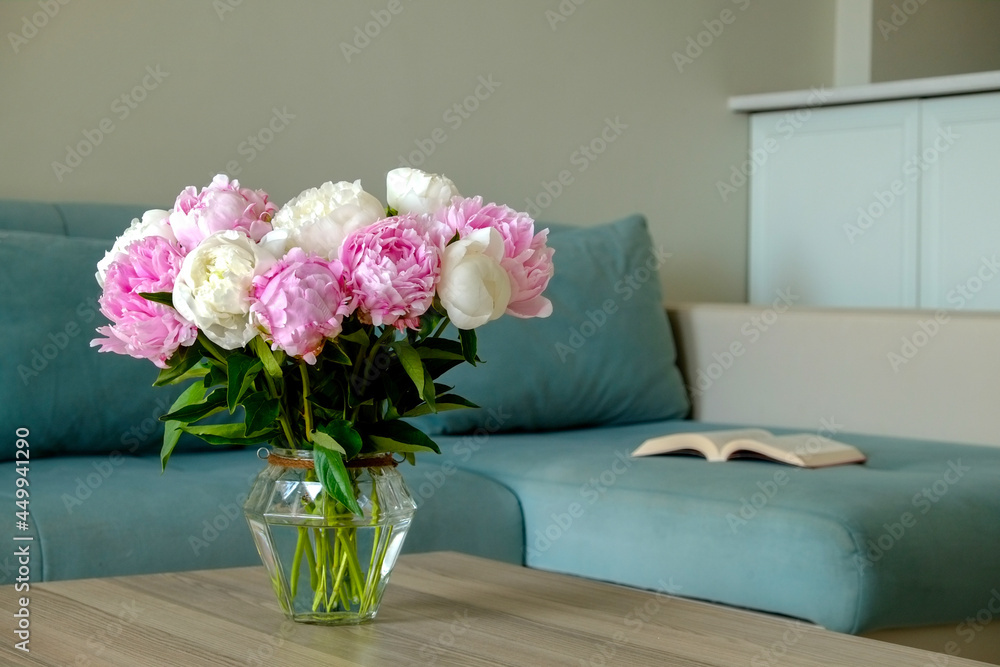Close up shot of wooden coffee table with bouquet of beautiful white peony flowers in glass vase on foreground and blue textile couch on the background. Copy space for text, close up. Close up shot of wooden coffee table with bouquet of beautiful white peony flowers in glass vase on foreground and blue textile couch on the background. Copy space for text, close up.