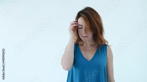Portrait of serious girl in blue dress and red hair thoughtfully touches her forehead and hair, turns her head to the side standing isolated over white wall background in the studio. Beauty concept