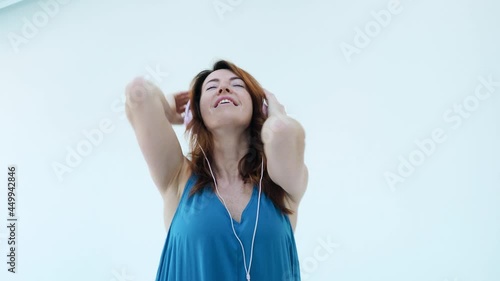 Close up. Positive young red hair girl with pink headphones smiling and dancing while listening music. She touches big headphones, enjoying songs and move to the music isolated on white studio wall