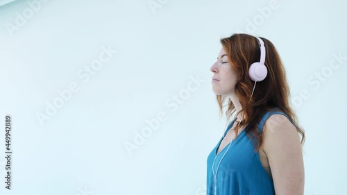 Close up. Serious red hair girl with pink headphones and blue dress listening music and dancing slightly with closed eyes standing sideways. Enjoying songs, meditation, time alone