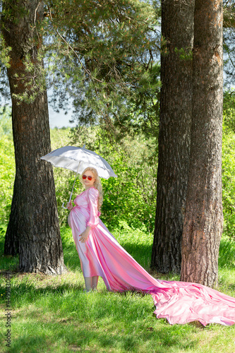 pregnant woman walks in the summer in the forest in a beautiful pink dress under a white umbrella in the sun goggles