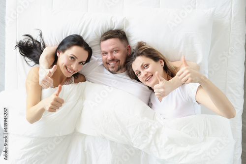 Two smiling women and man lie on bed and hold their thumbs up
