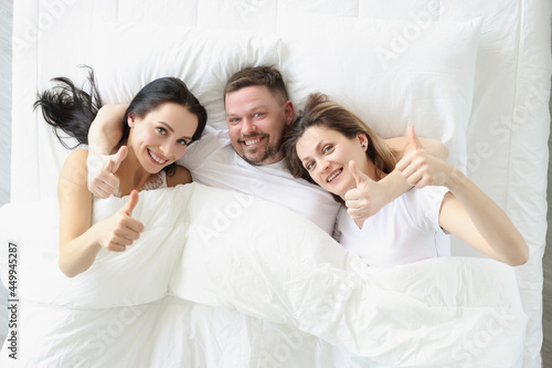 Принти на полотні Two smiling women and man lie on bed and hold their thumbs up