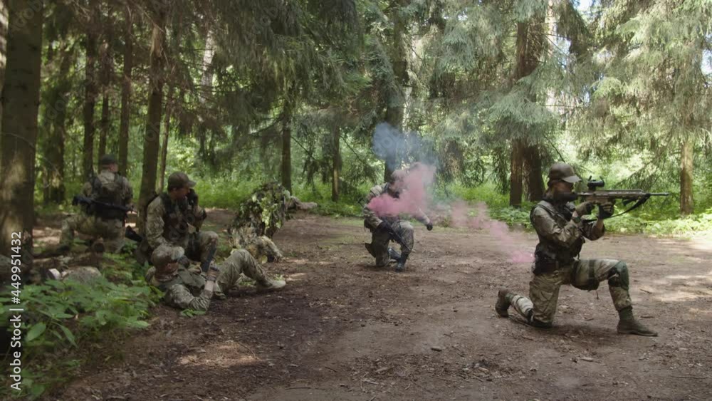 Fully equipped counter-terrorism group in camouflage holding assembly point in forest area. Military squad leader making radio call while soldier applying red hand flare evacuation signal Fully equipped counter-terrorism group in camouflage holding assembly point in forest area. Military squad leader making radio call while soldier applying red hand flare evacuation signal