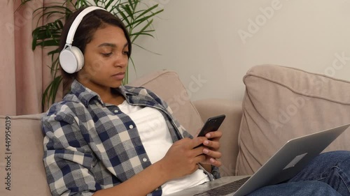 African-American woman with headphones listening to music lying at home on the couch with the phone in her hands. The concept of a holiday home.