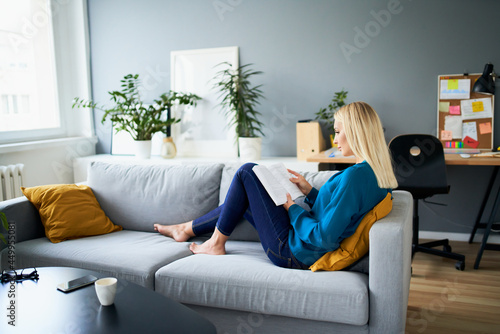 Young woman reading book sitting on the sofa at her apartment