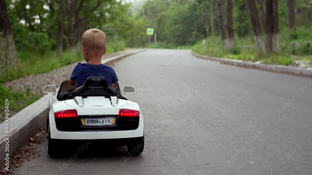Back of a 4 years old boy driving a white toy battery car in the park along the road. Summer activity with electric car in the park Back of a 4 years old boy driving a white toy battery car in the park along the road. Summer activity with electric car in the park