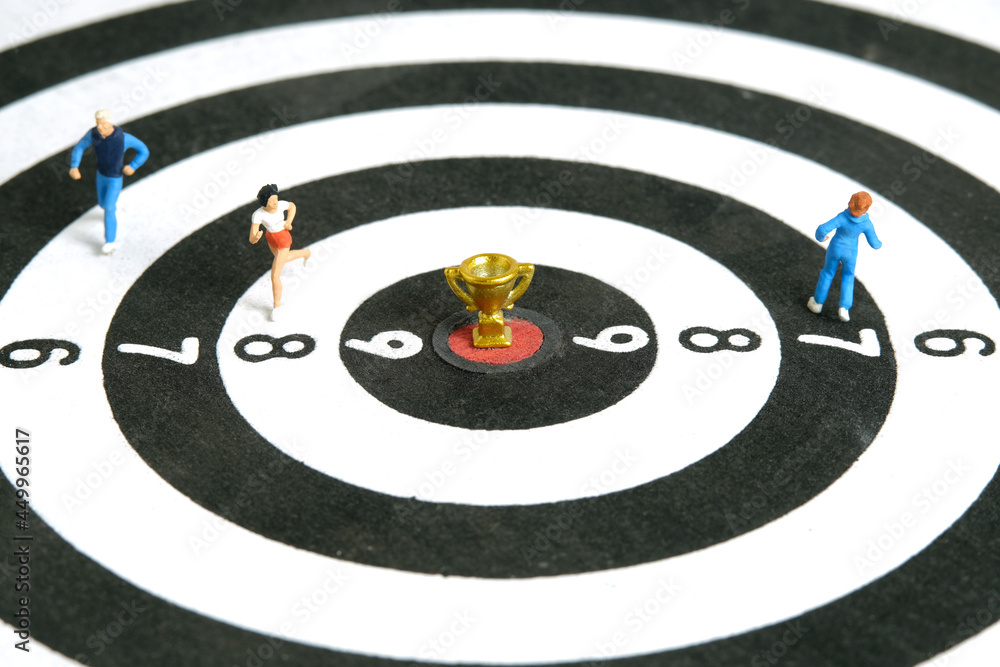 Miniature people toy figure photography. Winning a run competition concept. A runner running above dartboard with golden trophy at the center, isolated on white background Miniature people toy figure photography. Winning a run competition concept. A runner running above dartboard with golden trophy at the center, isolated on white background