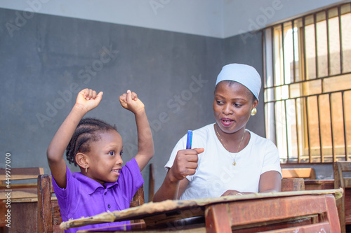 African Nigerian mother or teacher sitting together with her girl child in a classroom, helping her with her studies towards excellence in her, school, education and career
