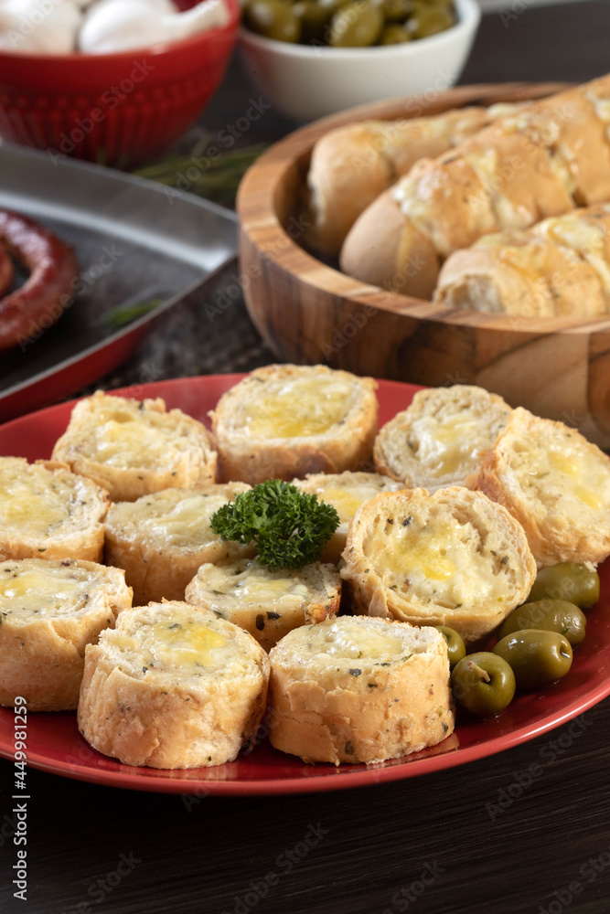 Garlic bread on a red plate on the barbecue table with sausage, cheese, rosemary, olives and cherry tomatoes.