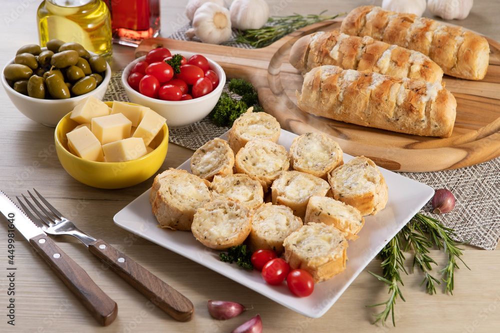 Garlic bread in white square plate on the table with cheese, rosemary, olives and cherry tomatoes. Top view. Garlic bread in white square plate on the table with cheese, rosemary, olives and cherry tomatoes. Top view.