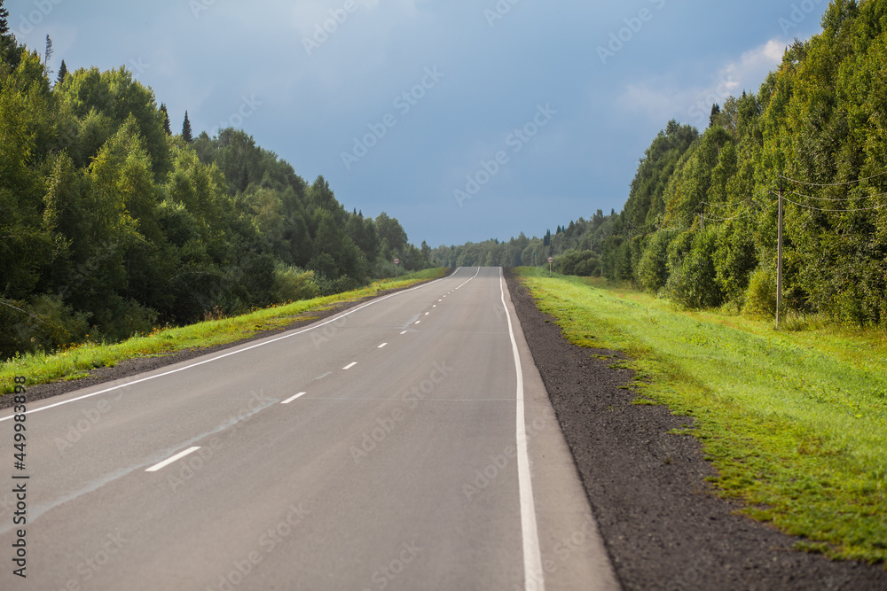 Straight road with a marking on the nature background. Open Road in future, no cars, auto on asphalt road through green forest, trees. Clouds on blue sky in summer, sunshine, sunny day. Bottom view Straight road with a marking on the nature background. Open Road in future, no cars, auto on asphalt road through green forest, trees. Clouds on blue sky in summer, sunshine, sunny day. Bottom view