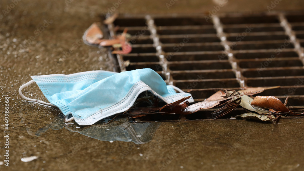 A close up of a discarded used personal protective face mask entering a metal grate on a drain. Growing environmental concern regarding safe disposal of millions of face masks.
