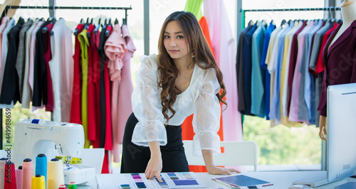 Cute and enerfyful female Asian designer working at desk in design room among clothes and fabric color sample