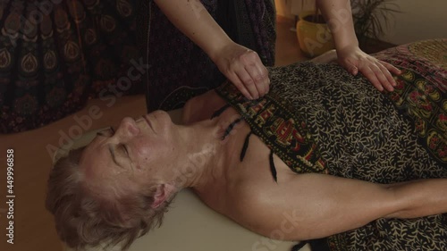 An older woman relaxing on her back on bed while a professional masseuse is massaging her chest with hands. A professional masseuse lady massaging her customer 's chest and abdomen using hands. photo