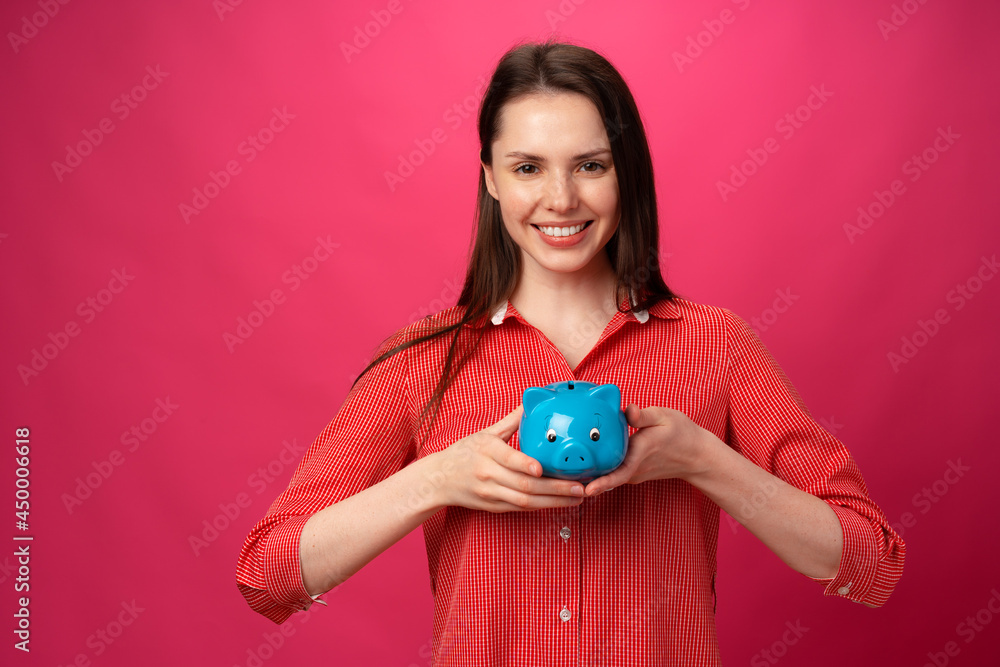 Happy smiling young woman holding blue piggy bank against pink background Happy smiling young woman holding blue piggy bank against pink background