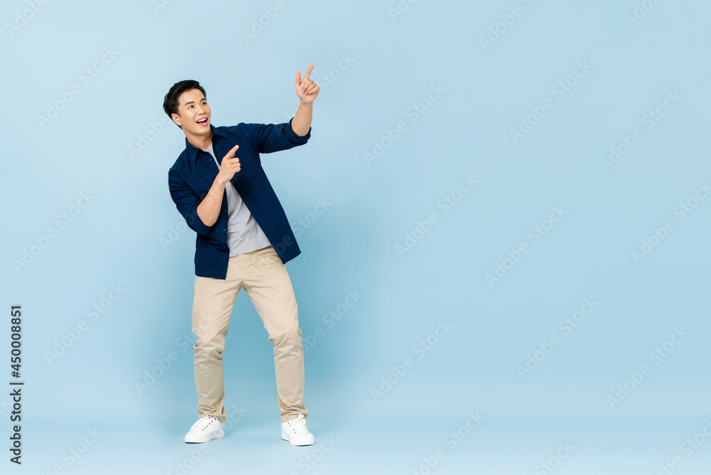Full length portrait of smiling young handsome Asian man pointing fingers to empty space aside in isolated studio light blue background Full length portrait of smiling young handsome Asian man pointing fingers to empty space aside in isolated studio light blue background