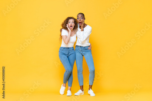 Full length portrait of shocked interracial millennial woman friends gasping with hands cupping mouths in isolated studio yellow color background
