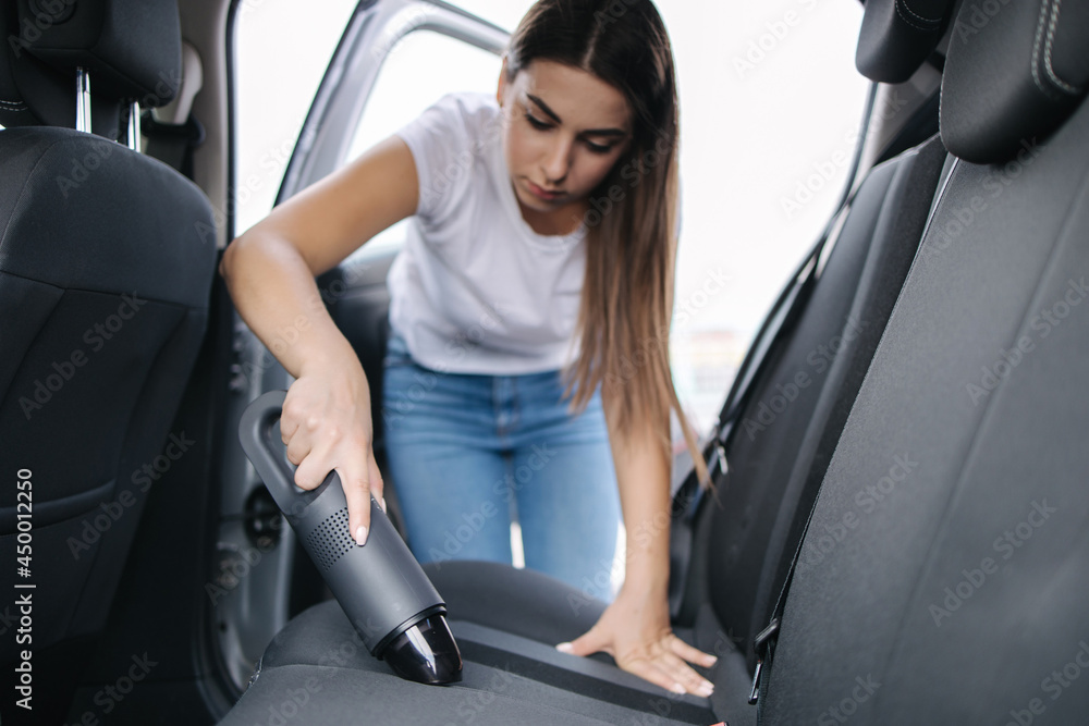 Woman vacuuming her car in the garage at home. Female using portable vacuum cleaner to remove dust and dirt. Car interior cleaning Woman vacuuming her car in the garage at home. Female using portable vacuum cleaner to remove dust and dirt. Car interior cleaning
