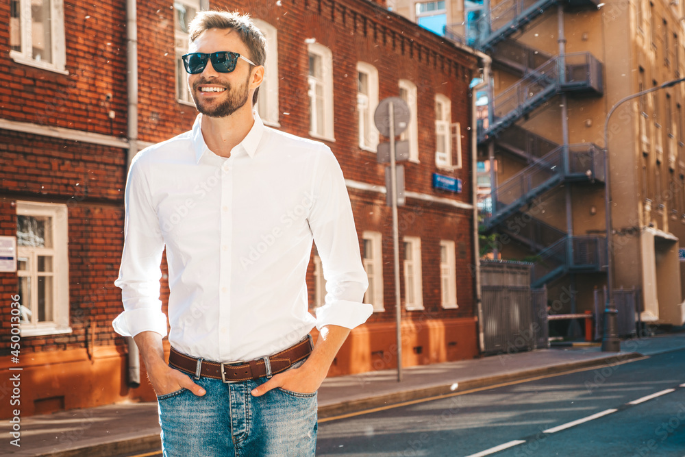 Fototapeta premium Portrait of handsome confident stylish hipster lambersexual model.Modern man dressed in white shirt. Fashion male posing in the street background near skyscrapers in sunglasses